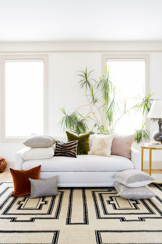 A white couch is filled with decorative pillows of all shapes sizes and colors. The pillows are brown, olive, cream, blush, and grey. The rug is a tan color with a black geometric symmetrical design.