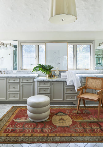 A spacious powder room with large white countertops and grey cabinets. In the center is a large red rug with yellow and green detailing. On top of the rug is a round footstool and yellow wicker chair.