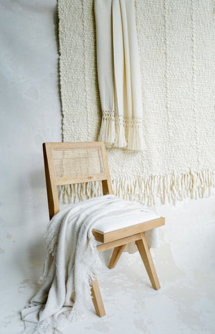 In the center is a wicker chair with a oak frame and a white linen cushion. Draped on the chair is a chunky white knit throw blanket. On the wall behind the chair are two cream colored blankets hanging like a tapestry.