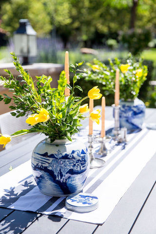 There are two blue and white vases with yellow tulips sitting on an outdoor table. Between the vases are four yellow candlesticks. Under the vases is a white linen runner.