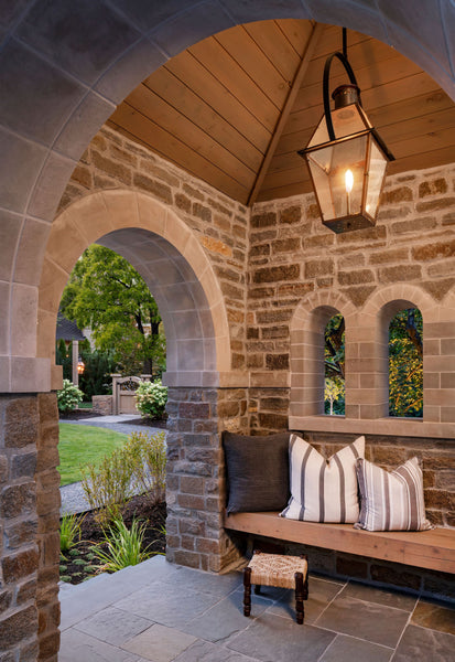 It is an enclosed patio with brick archways and slate flooring. A warm lantern hangs from the ceiling. On the back wall is a wooden built in bench with three pillows.