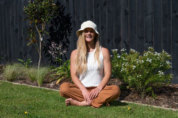 Caliwoods founder, Shay Lawerence, a caucasian women in her 30's, sitting on lawn in front of a flower garden wearing a white bucket hat, singlet and pants.