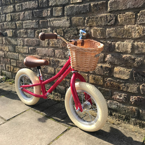 A Red Gingersnap Balance Bike with wicker basket leaned against a brown brick wall 