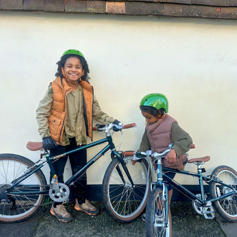 Tow children smiling while standing with their bikes against a white wall