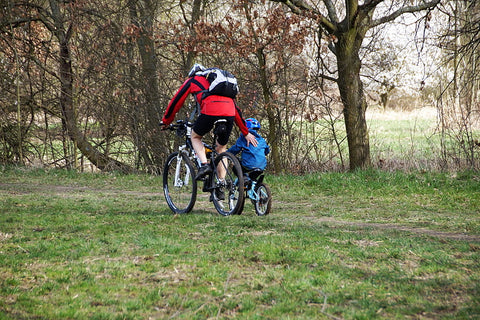 The parent guides the toddler's small bike along a forest trail.