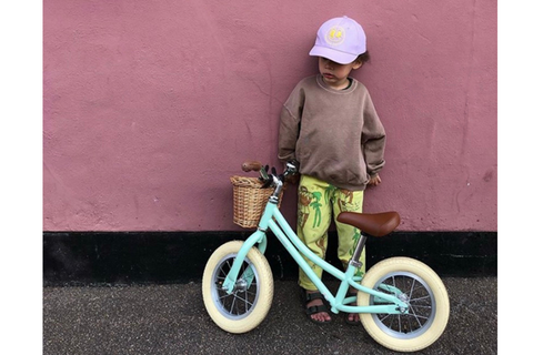 Boy with cap on standing against a pink wall next to a light blue balance bike