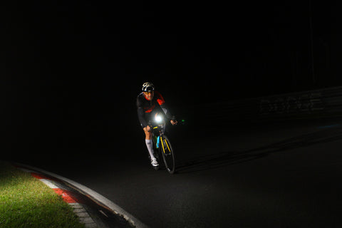 A man cycling on a road at night with illuminated bike lights.]