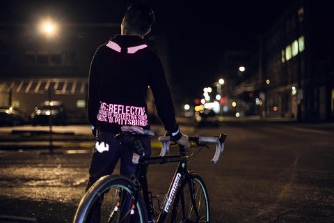 A cyclist donning a reflective jacket stands beside their bike at night.