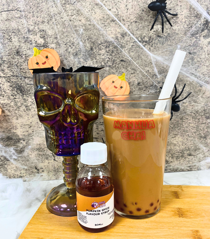 Marble background with spiderweb deocration, with black spides. In front glass of bubble tea with a skull decorative glass to the left and a bottle of pumpkin spice syrup in front.