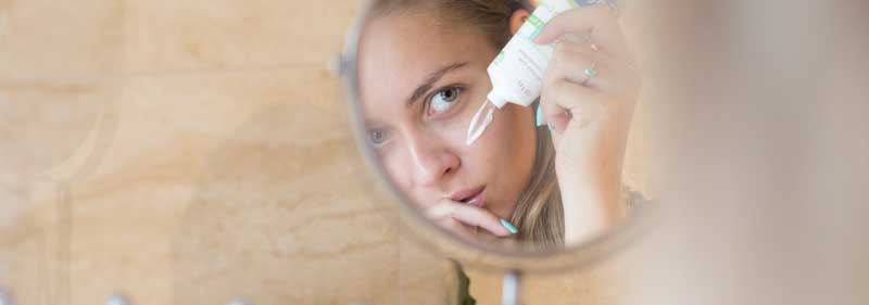 Reflection of young woman in small mirror putting face cream