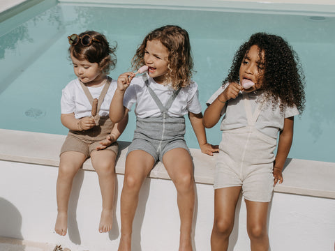Three kids sitting by a pool and eating an ice-cream during summer time. They are wearing the Silly Silas cotton shorties in peanut bland, in blue and in cream.