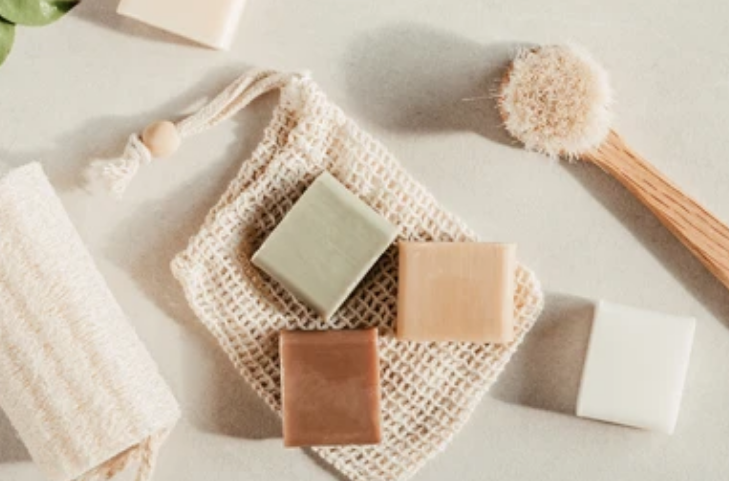 Three handmade soaps placed on a cream surface with brush and a pink towel.