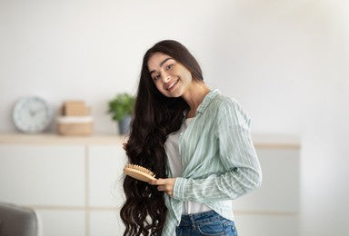 Portrait of lovely Indian lady brushing her wavy long hair, using wooden brush indoors. Cheerful young woman taking care of herself at home. Domestic spa salon, wellness and beauty concept