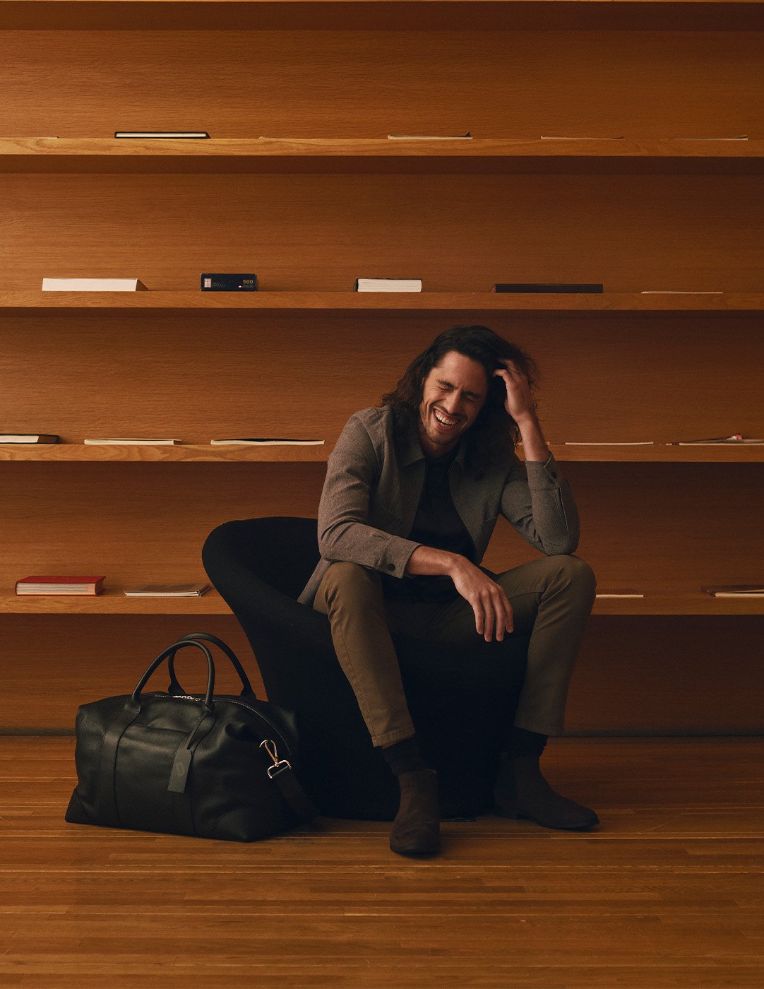 Man sitting in front of book shelves smiling with duffle bag at his feet