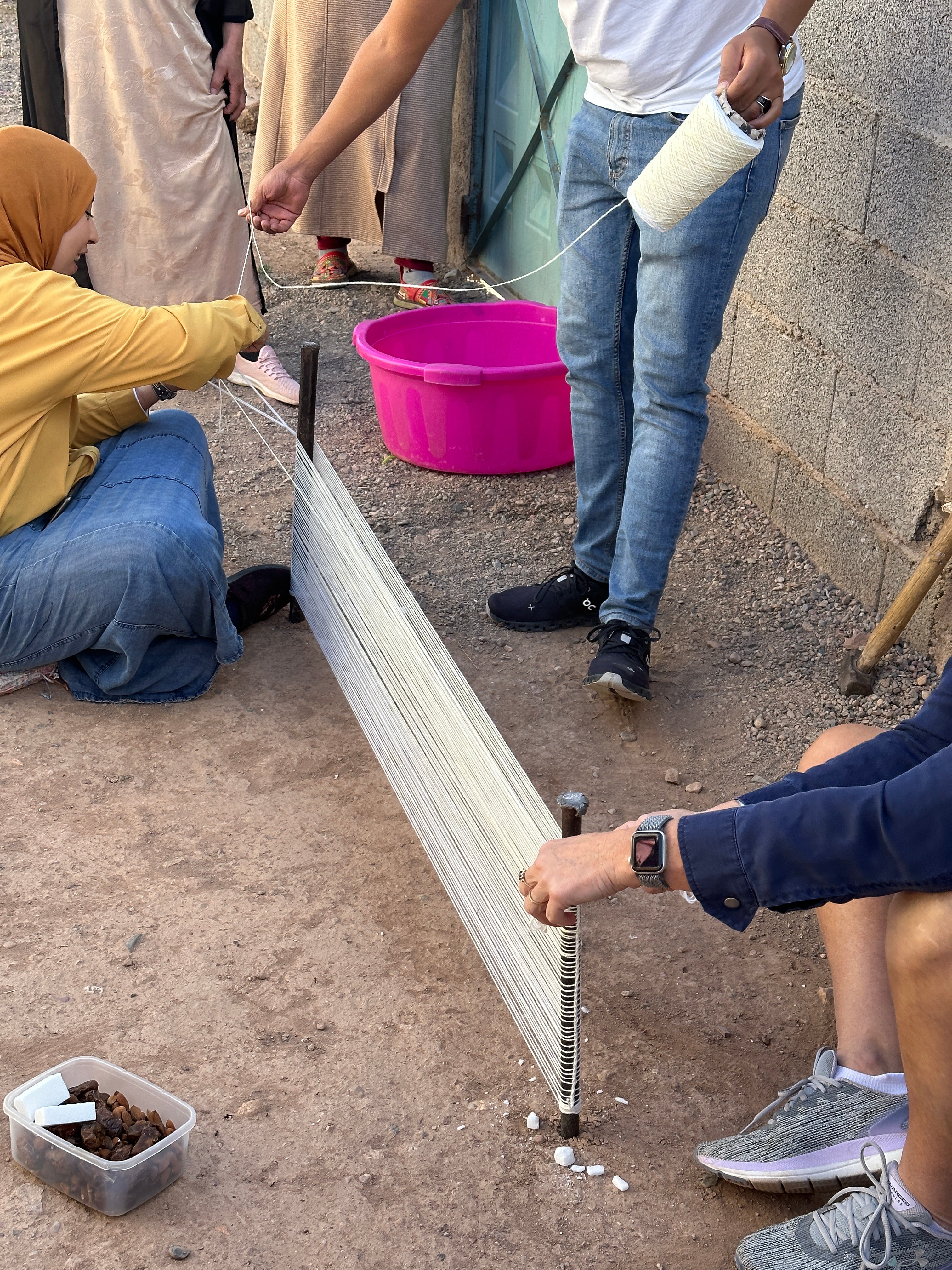 A group of Moroccan community members work together to wind a warp for weaving around metal stakes in the ground.