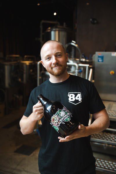 Photo: Nick Glynn standing in front of brewing system holding Waterloo Brewing branded growler featuring art of a hummingbird and flowers