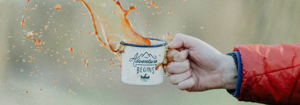 Hand holding a splashing coffee cup in nature that says "the adventure begins".
