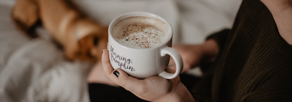 Woman holding a cup of coffee on a bed with a dog in the background. Coffee mug says "morning, pumpkin" on the side.