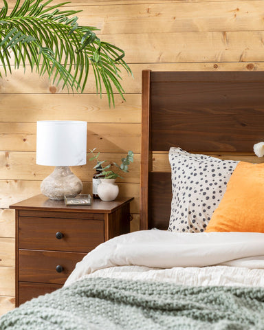Solid wood headboard and nightstand in a bedroom with a neutral bedspread and palm leaf in the background. 