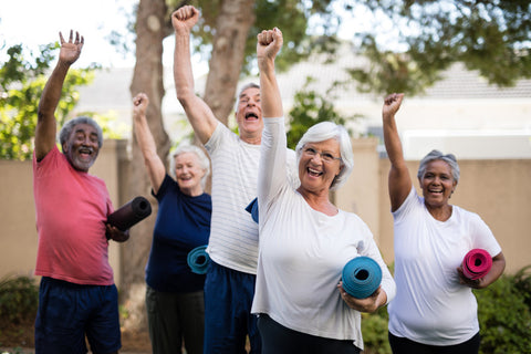 A group of enthusiastic seniors raising their hands while holding their yoga mats.