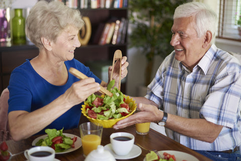 A senior couple smiling and looking at each other while enjoying their salad meal paired with coffee and juice.