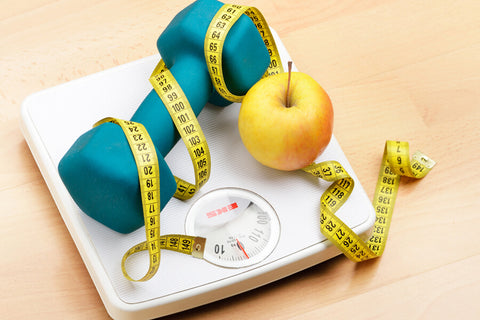 A weighing scale with an apple, a blue dumbbell, and a tape measure on top