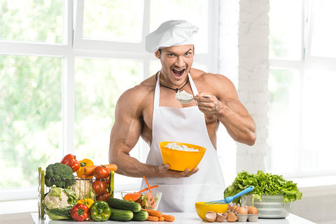 A male chef in white apron poses with healthy foods in his kitchen