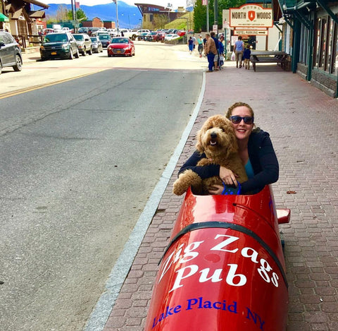 Top 10 Pet-Friendly Travel Destinations in the US | A dark colored goldendoodle sits in a bright colored bobsled with a female owner in the middle of Lake Placid sidewalk | Bubu Brands