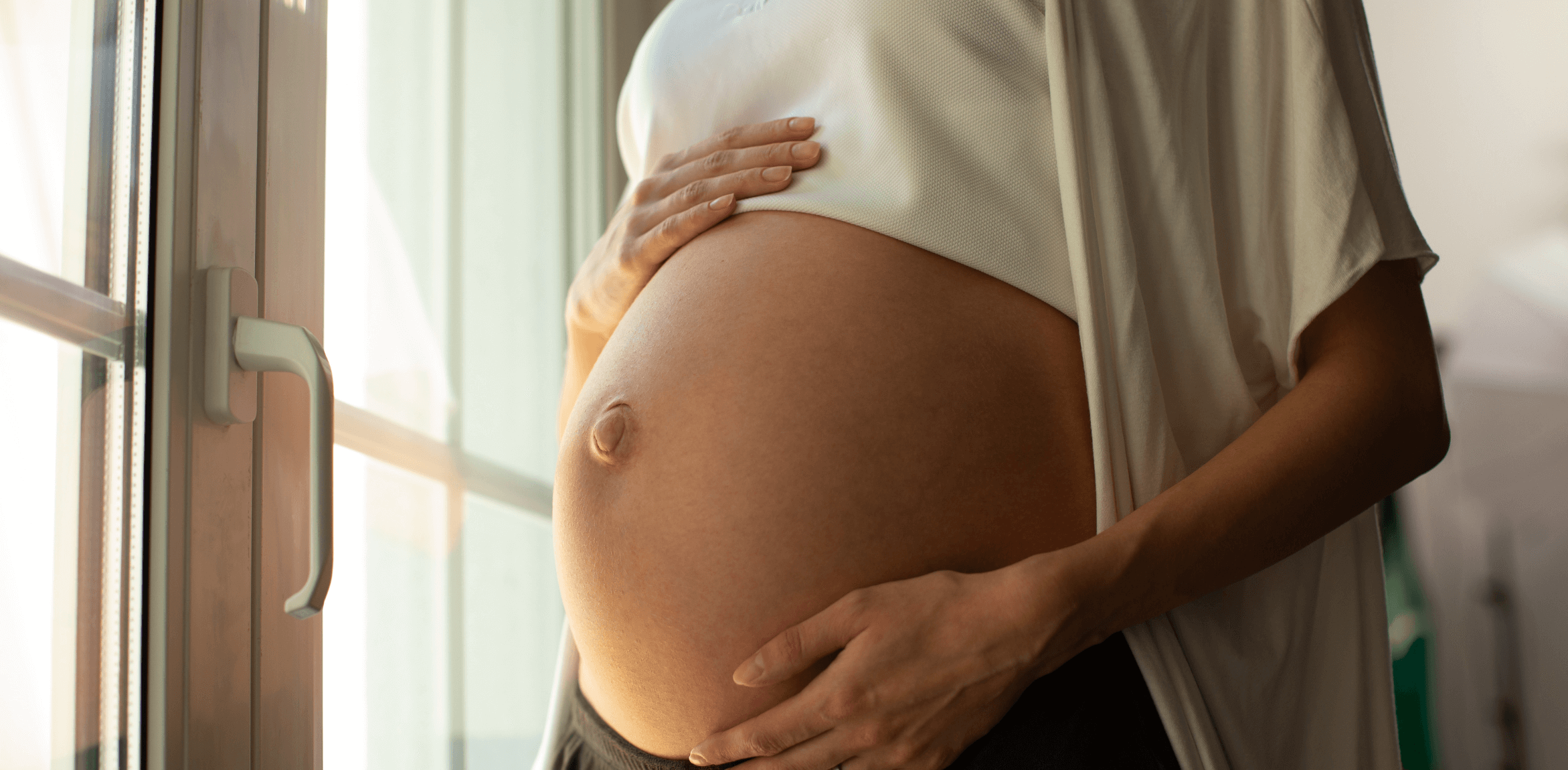 woman holding her bump near a window with image cut off so only her torso is in the frame