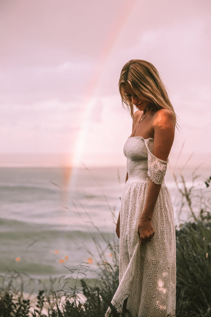 Side view of a woman wearing white see-through off-shoulder maxi dress standing on a grass field with rainbow behind