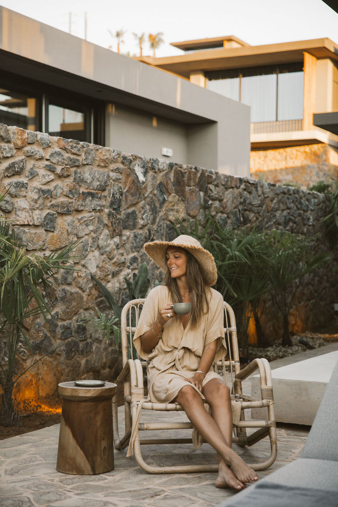 A woman looking beside her sitting on a beach chair wearing a loose v-neck tan dress while holding her coffee mug