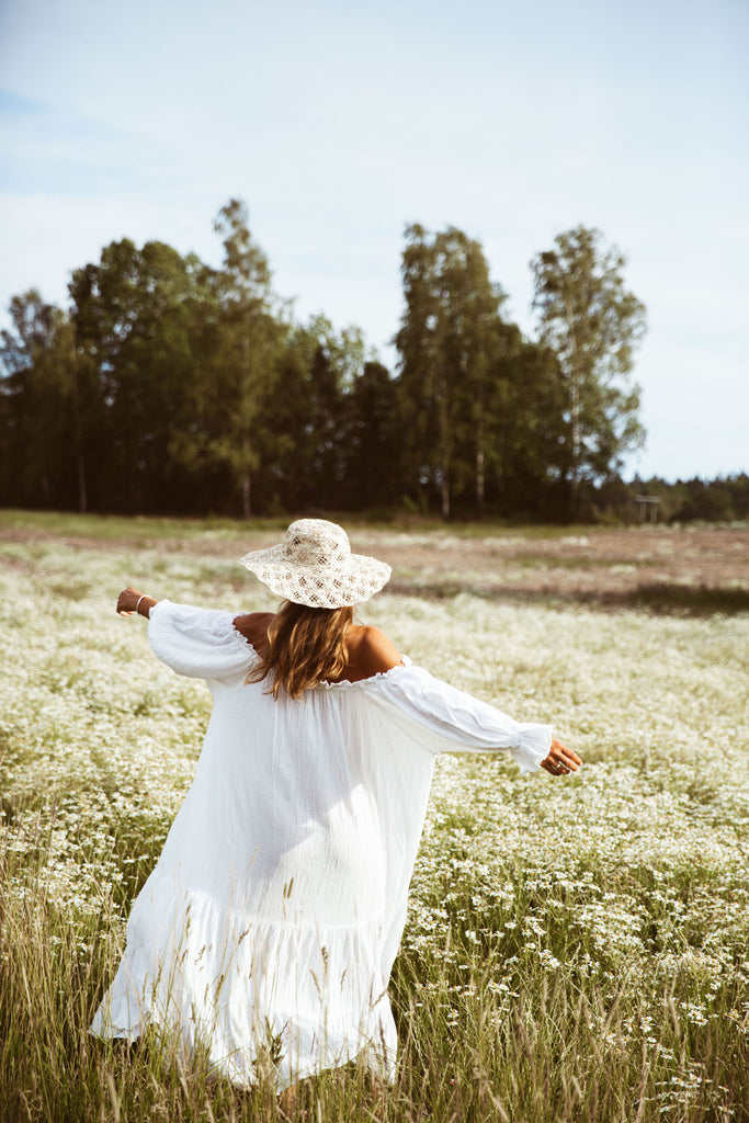 Back view of model Clara wearing a white off-shoulder maxi dress and a straw hat in a carefree pose on a field of tall grass