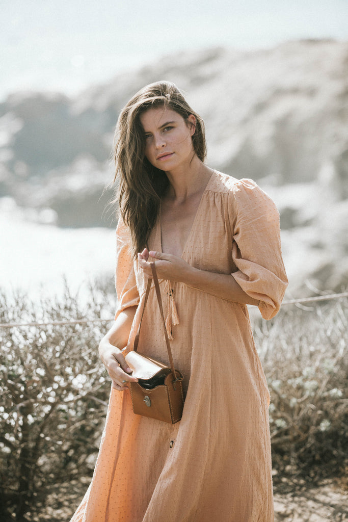 Our model Becca wearing a brown V-neck dress while holding her bag while looking at the camera