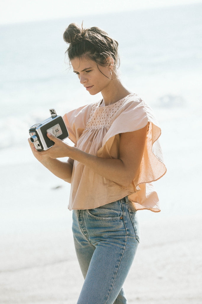 our model Becca, wearing light brown top and jeans at the beach while holding a video camera