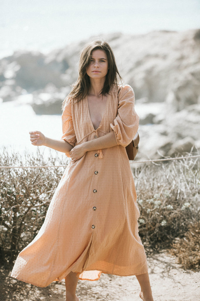 Our model, Becca, is wearing a brown V-neck dress with a bag on her shoulder while holding her waist.