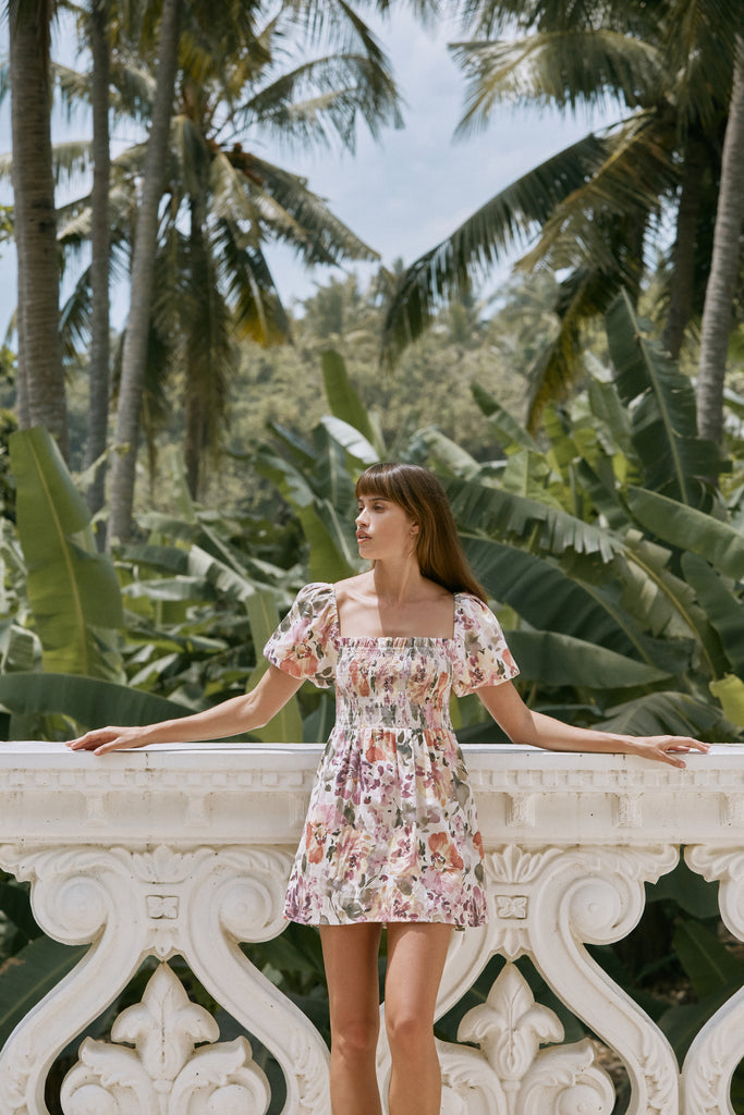 A woman wearing Camille Mini Dress leaning on a white balustrade with banana trees in the background