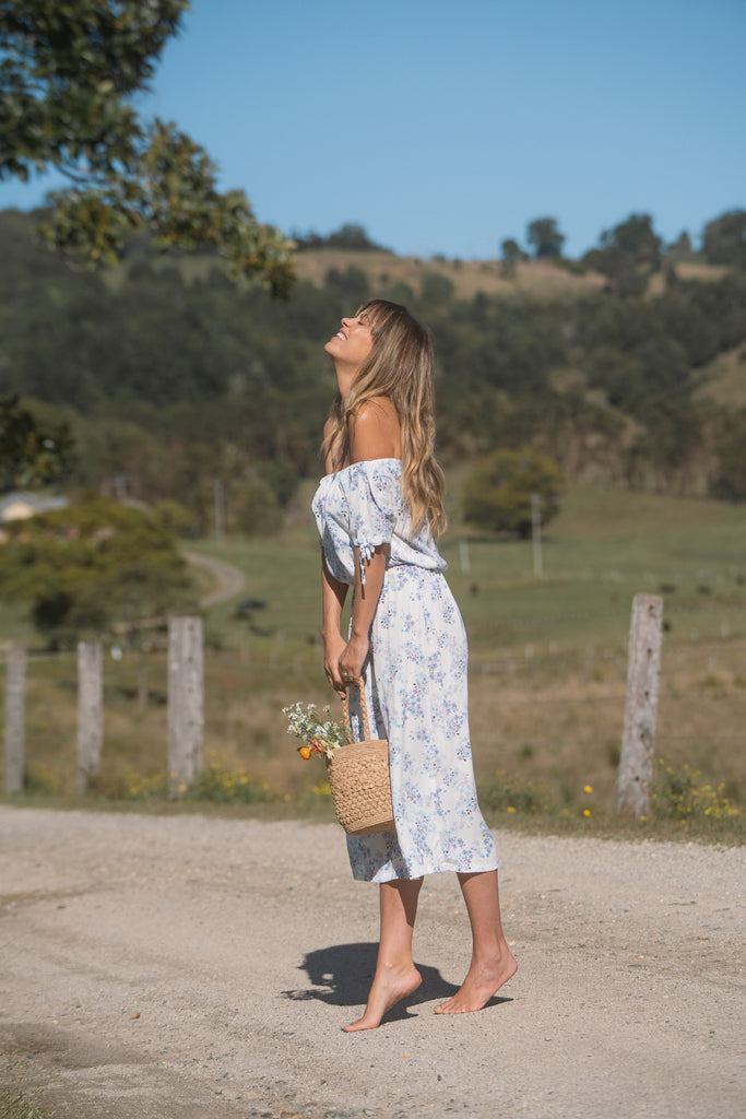 A side view of a woman wearing off-shoulder floral dress tip-toed on a dirt road holding a basket of flowers