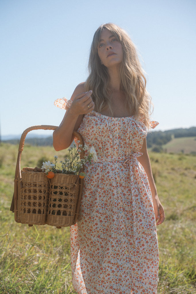 A woman wearing floral dress standing on a grass carrying a basket of flowers