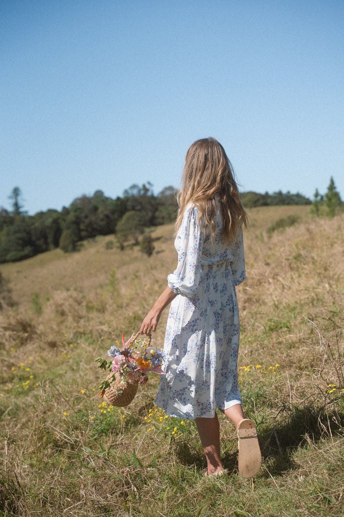 A woman wearing blue floral dress walking on a field of flowers while carrying a basket of flowers