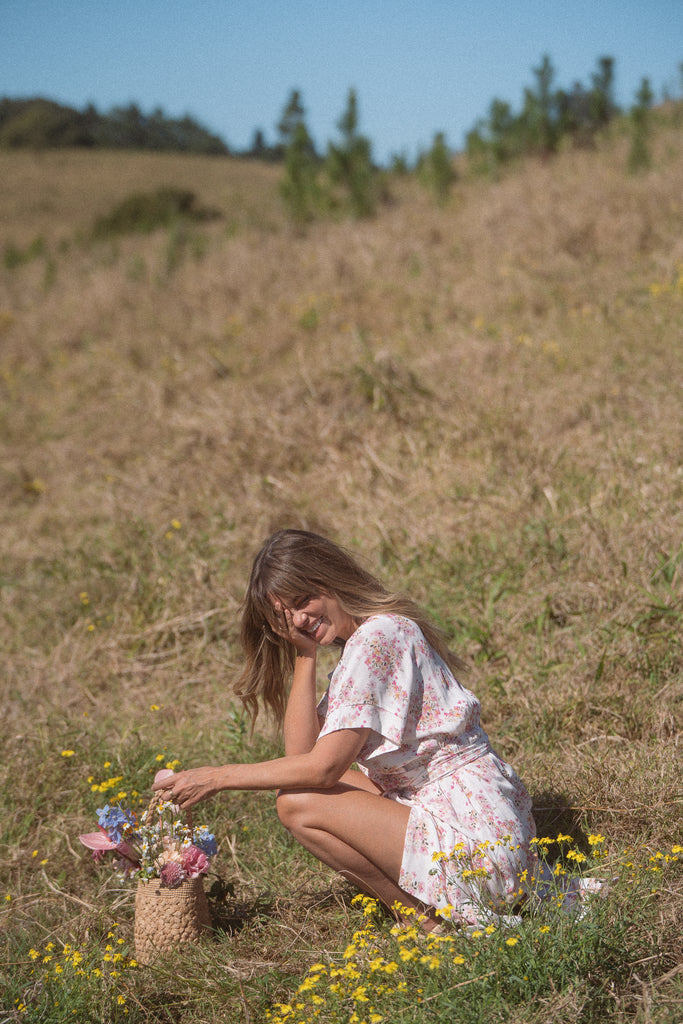 A woman wearing floral wrap dress sitting on a field of flowers while holding basket of flowers