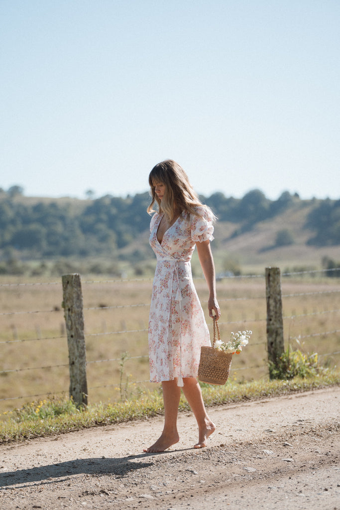 A woman wearing floral dress tip-toed on a dirt road while carrying a small basket with flowers