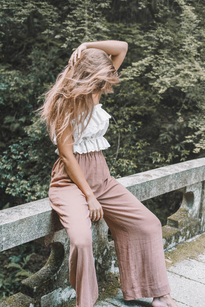 a woman wearing a ruffled tube top and rose gold trousers sitting on a stone bridge with her hands on her hair