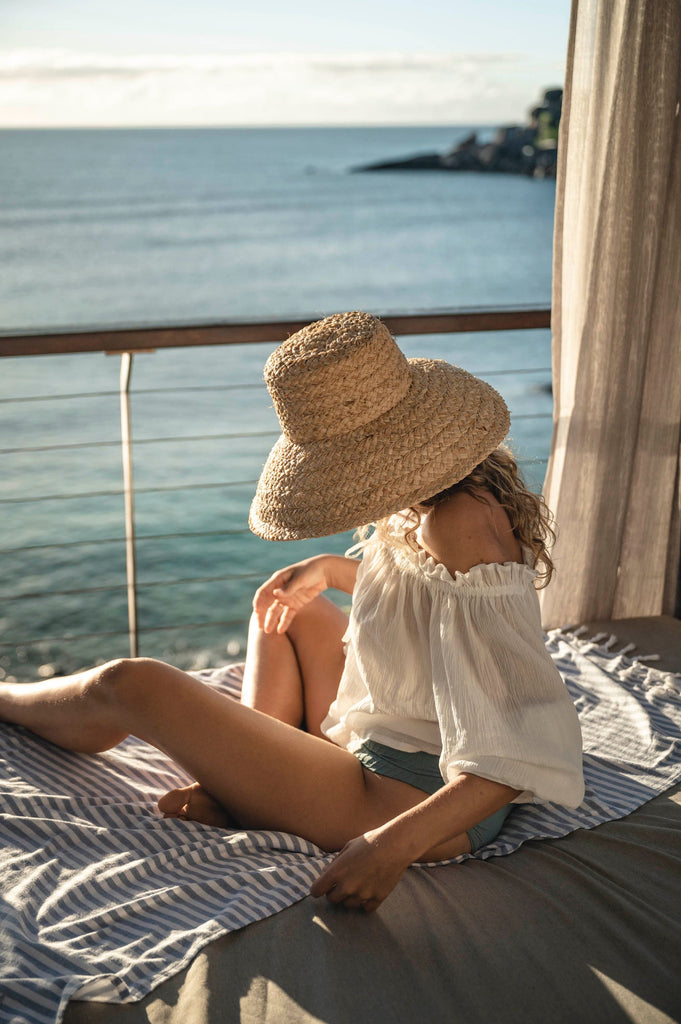 A woman sitting on a bed wearing an off-shoulder top and a gray underwear with a hat covering her face