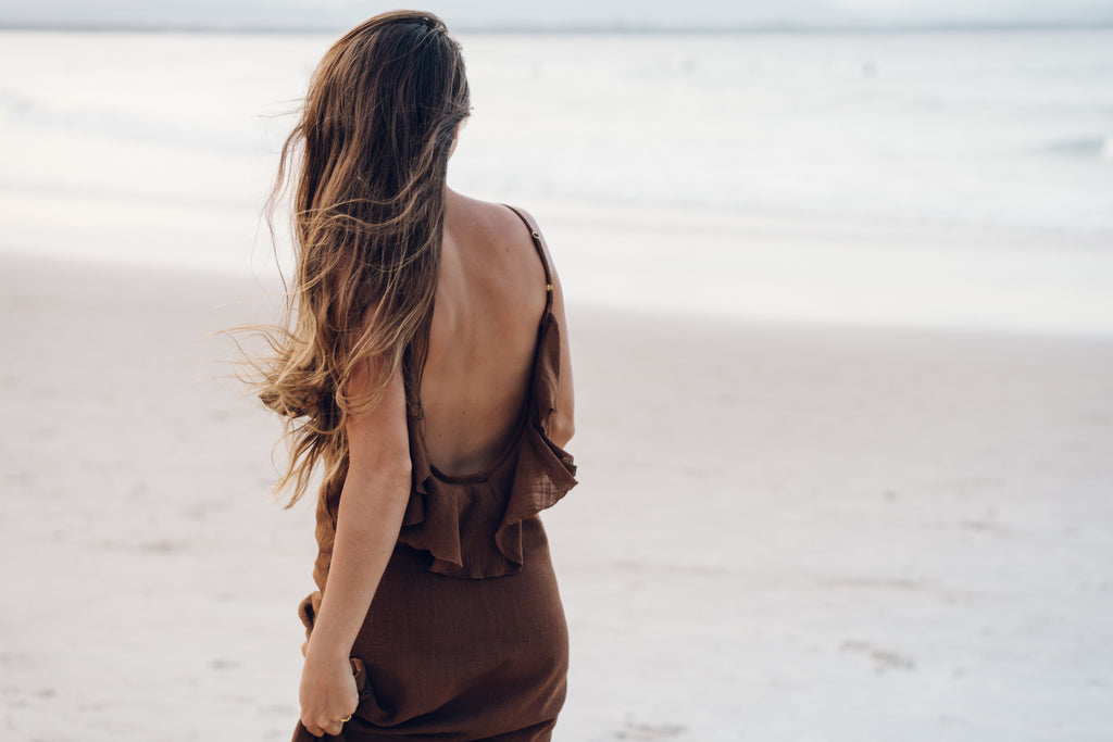 Back perspective of our model wearing dark brown sleeveless dress walking on the sea side