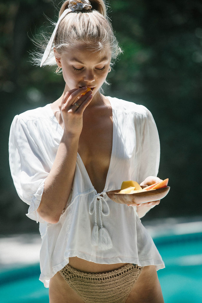 Our model Nina, wearing a white tie-front blouse and a light brown bikini while holding a bowl of bite-sized fruits