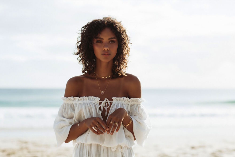 a woman looking at the camera in a white off shoulder see-through dress standing on the beach