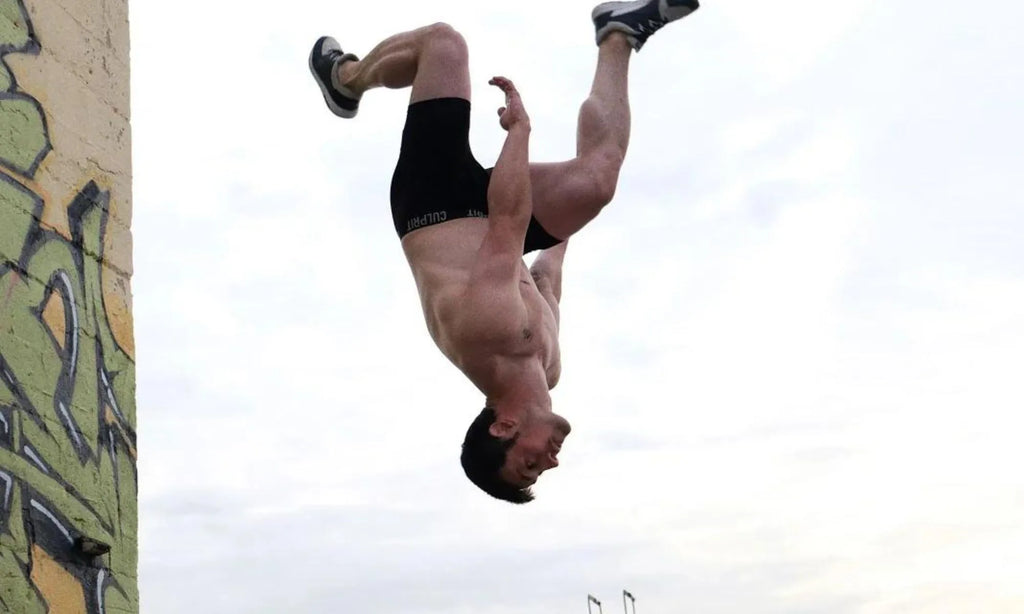 Shirtless man performing a flip in mid-air, wearing Culprit underwear and sneakers.
