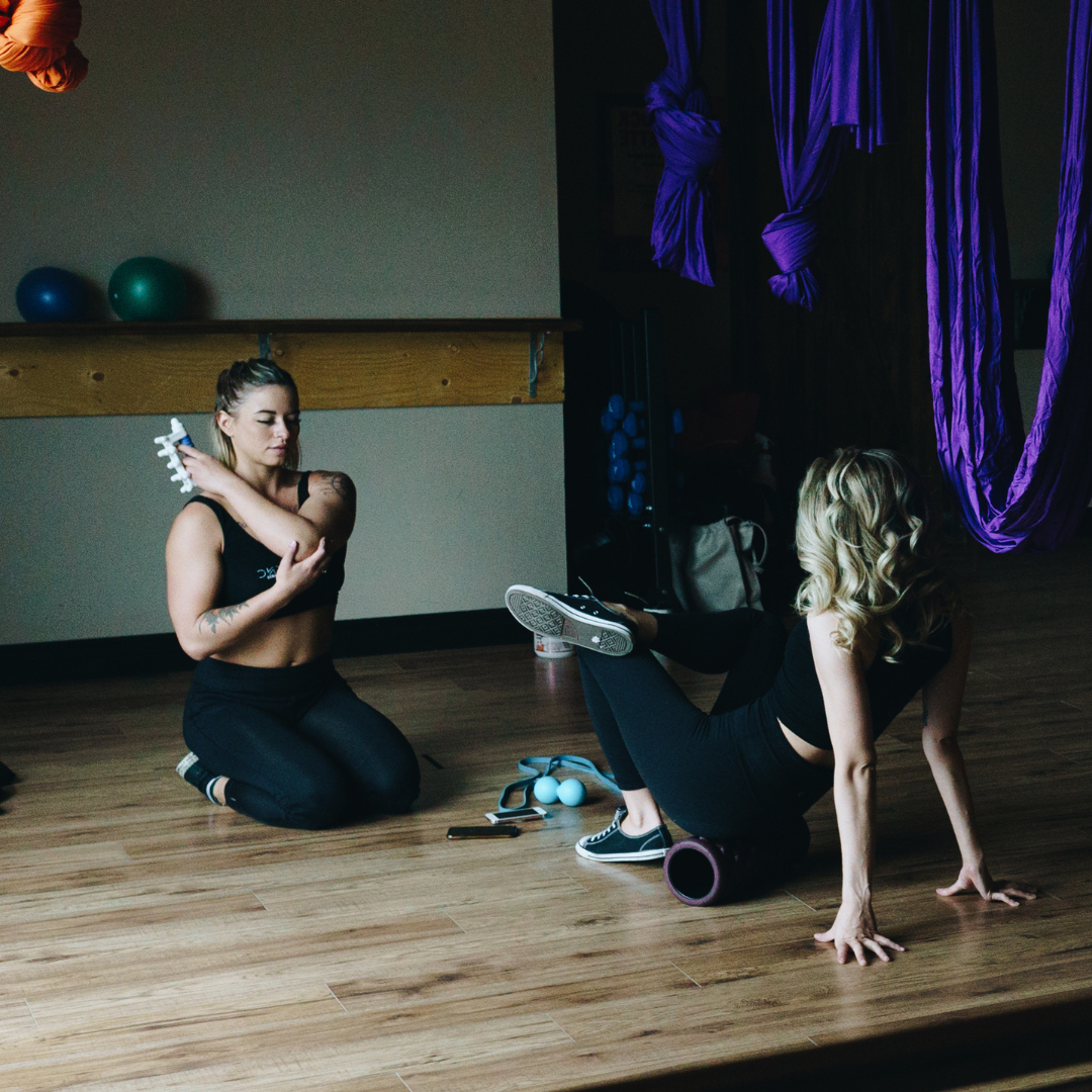 2 women in a gym setting using recovery tools, including the koa massage tool and foam roller