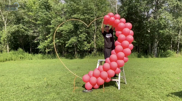 A person standing on a step ladder attaching red balloons to one side of a gold metal heart-shaped photo backdrop stand.