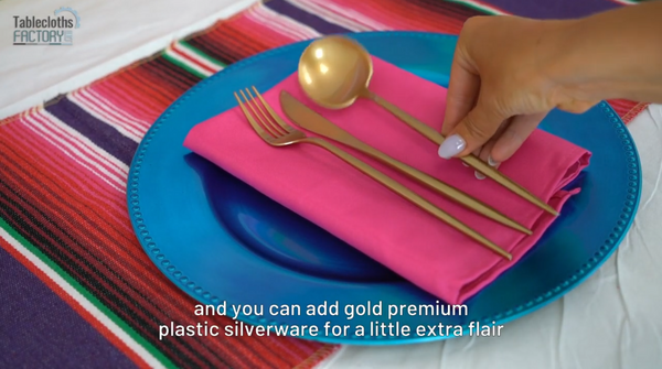 Setting a table with a blue charger plate, fuchsia napkin, and gold utensils for elegant fiesta-themed party decorations.